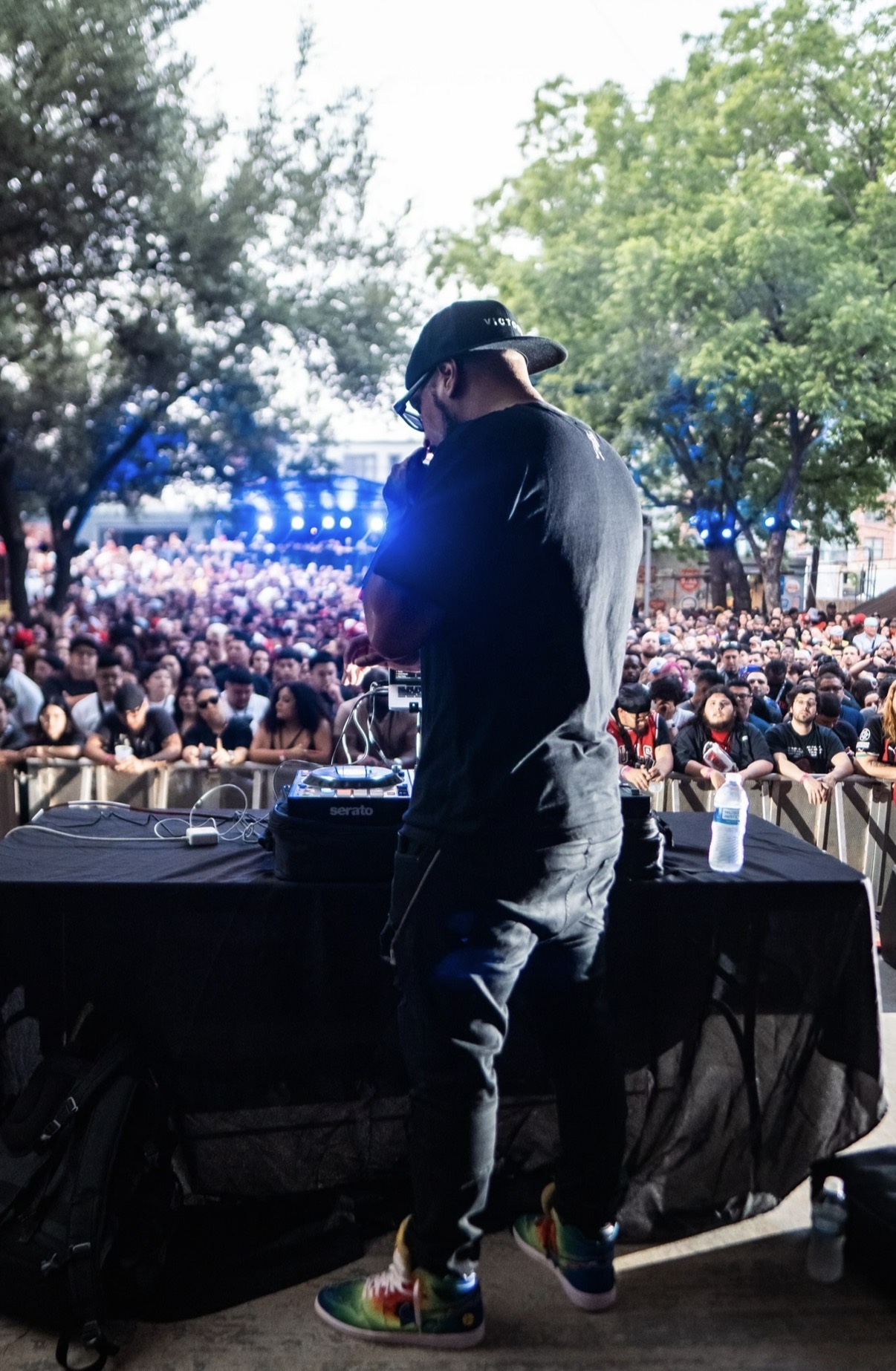 Person wearing a hat and dark clothing performing on stage with a large crowd and trees in background.
