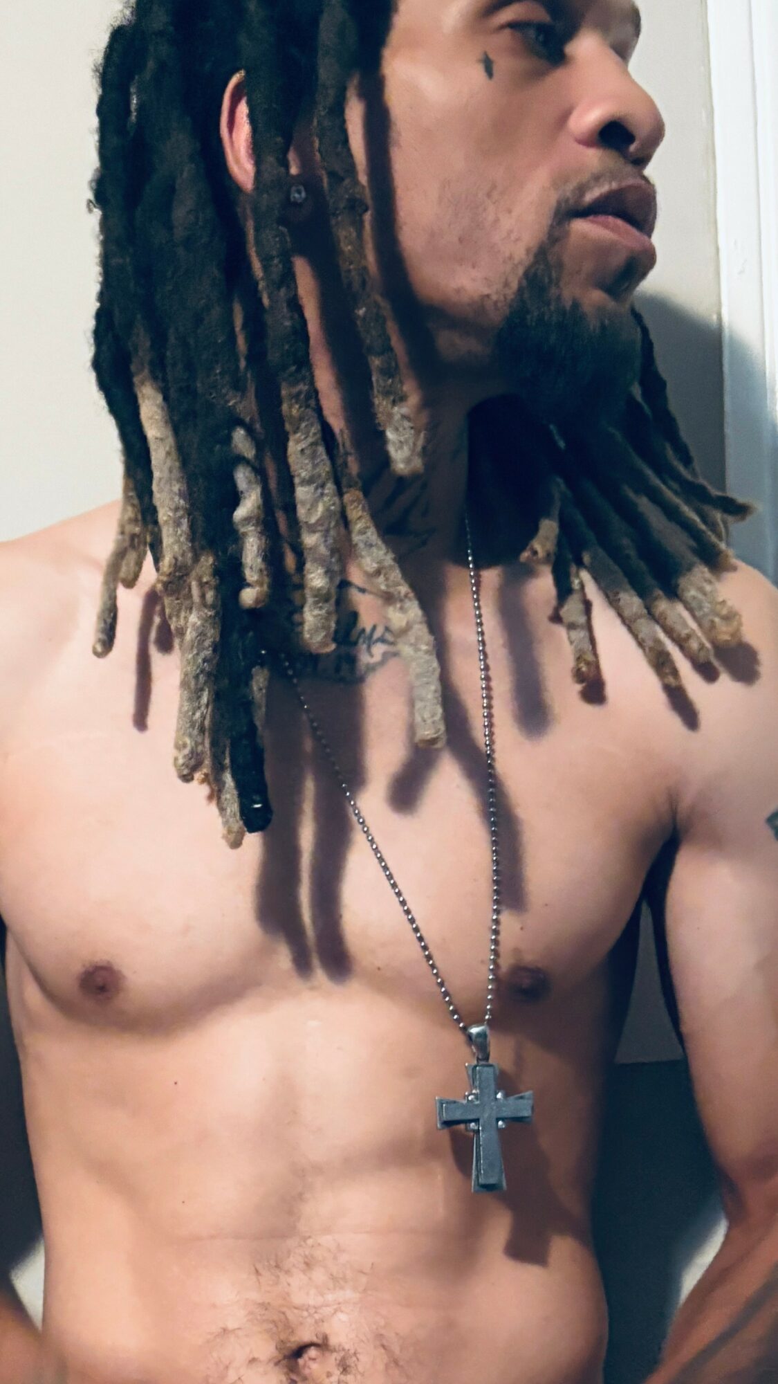 A shirtless man with dreadlocks wearing a cross necklace, looking to the side.