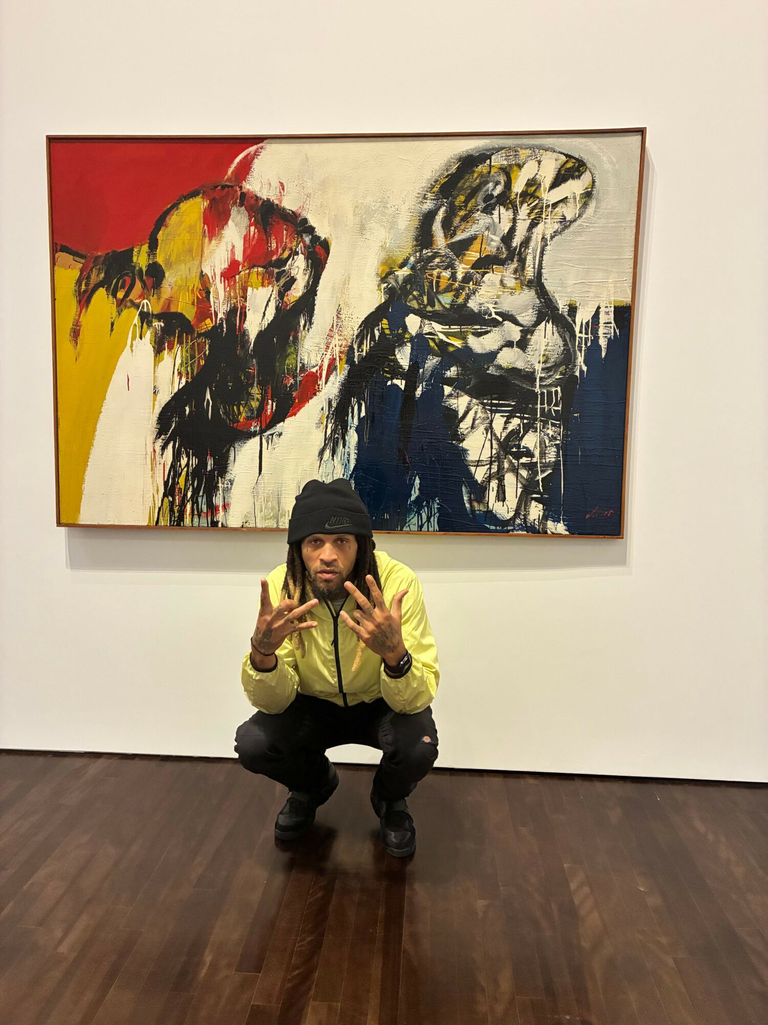 Person crouching next to colorful abstract painting on white wall, wooden floor, making peace sign with hand.