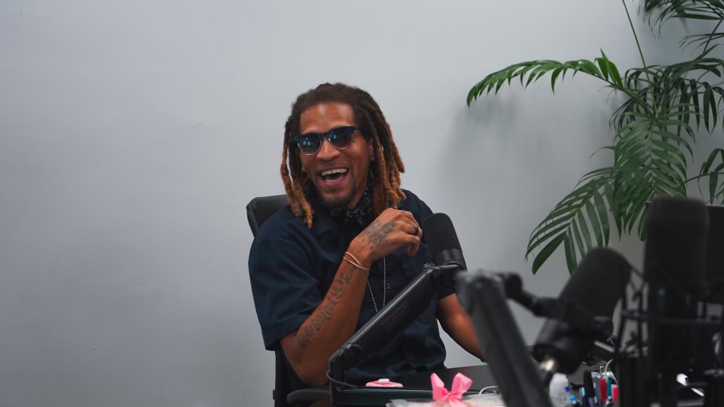 Person with dreadlocks and sunglasses smiling, sitting at a desk with a camera and pink flower, against a plain background.
