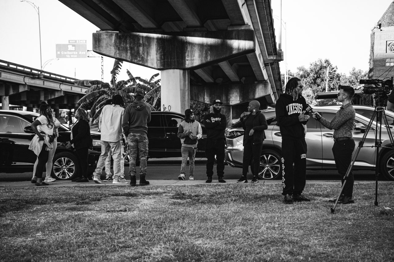 Group of people gathered outdoors under a bridge, some with cameras, in black and white.