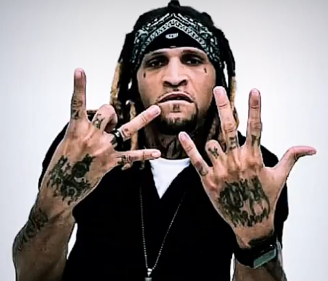Person with tattoos on hands and face, wearing a bandana, making hand gestures, against a plain background.