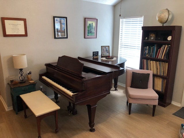 Room with a grand piano, chair, bookshelf, window, and framed pictures on the wall.