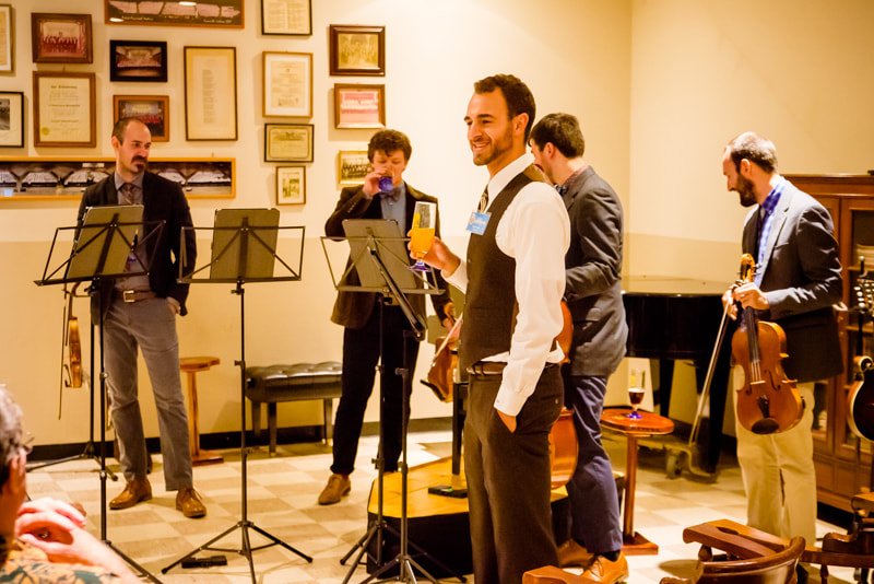 Group of musicians performing indoors with framed pictures on the wall, a man holding a drink, and a person playing guitar.