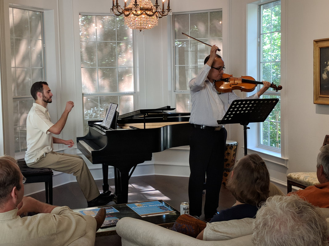 Two musicians perform in a bright room with large windows, one playing piano and the other playing violin, audience seated nearby.