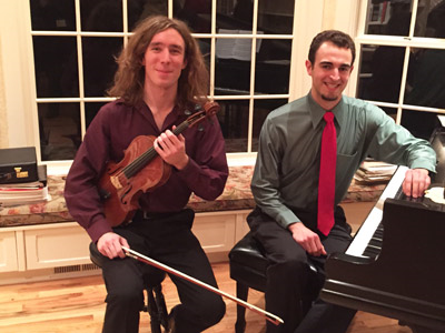Two men sitting indoors, one holding a violin and bow, the other at a piano, in front of large windows.
