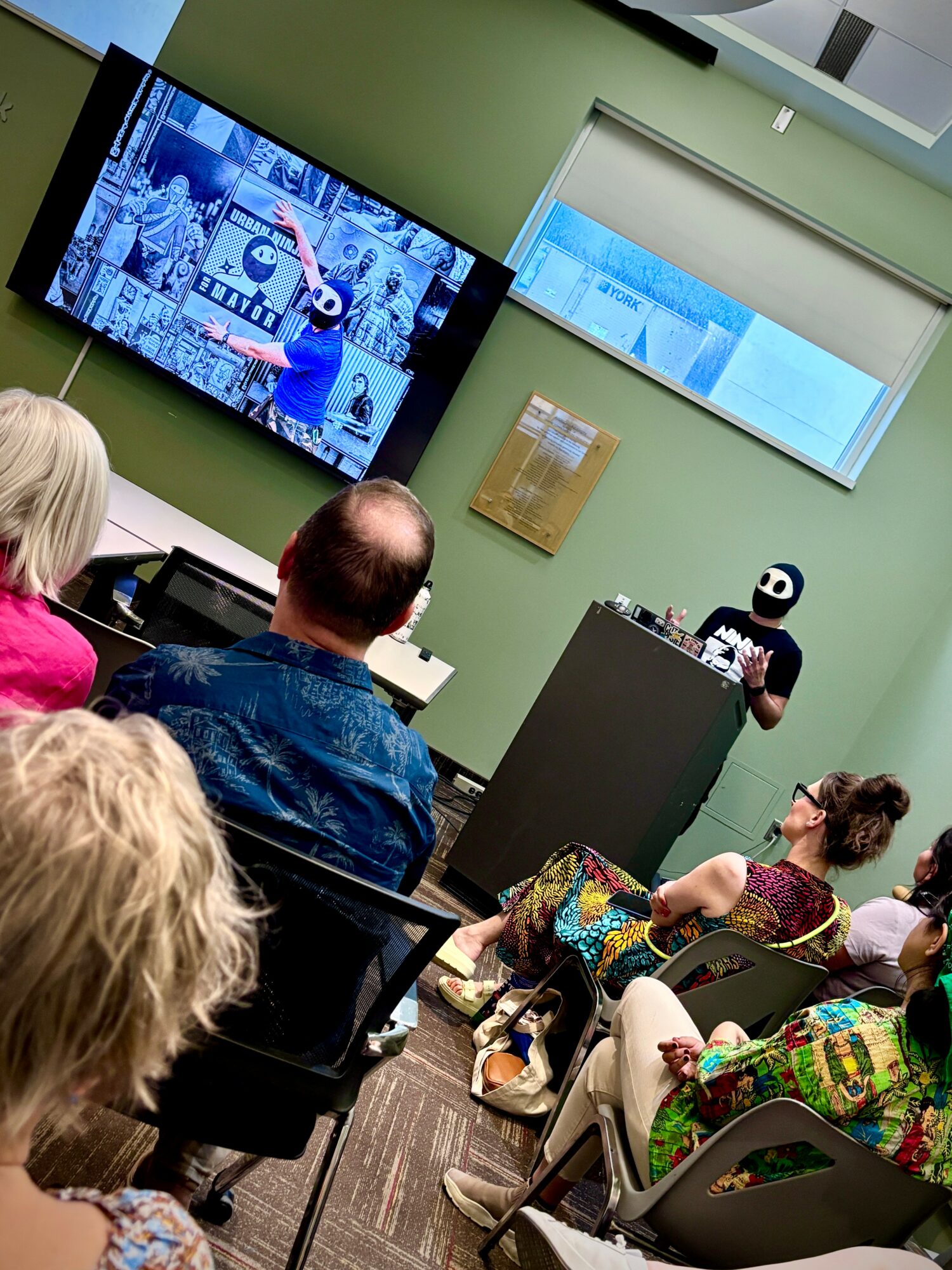 People seated in a room watching a presentation on a large screen, with a presenter at a podium and two screens on the wall.