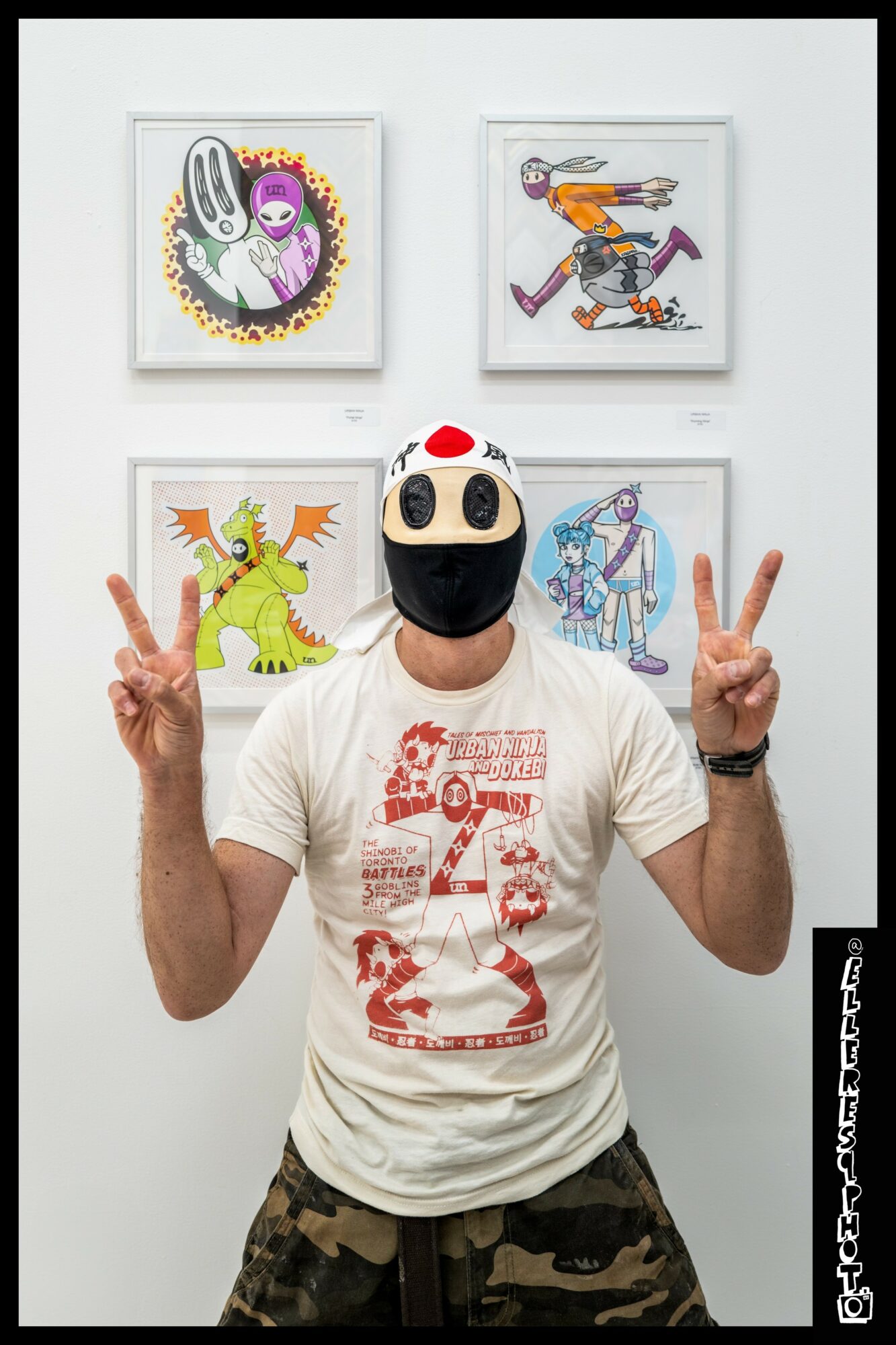 Person wearing a mask with large eyes and a white hat, making peace signs with both hands, in front of four framed colorful illustrations on a white wall.