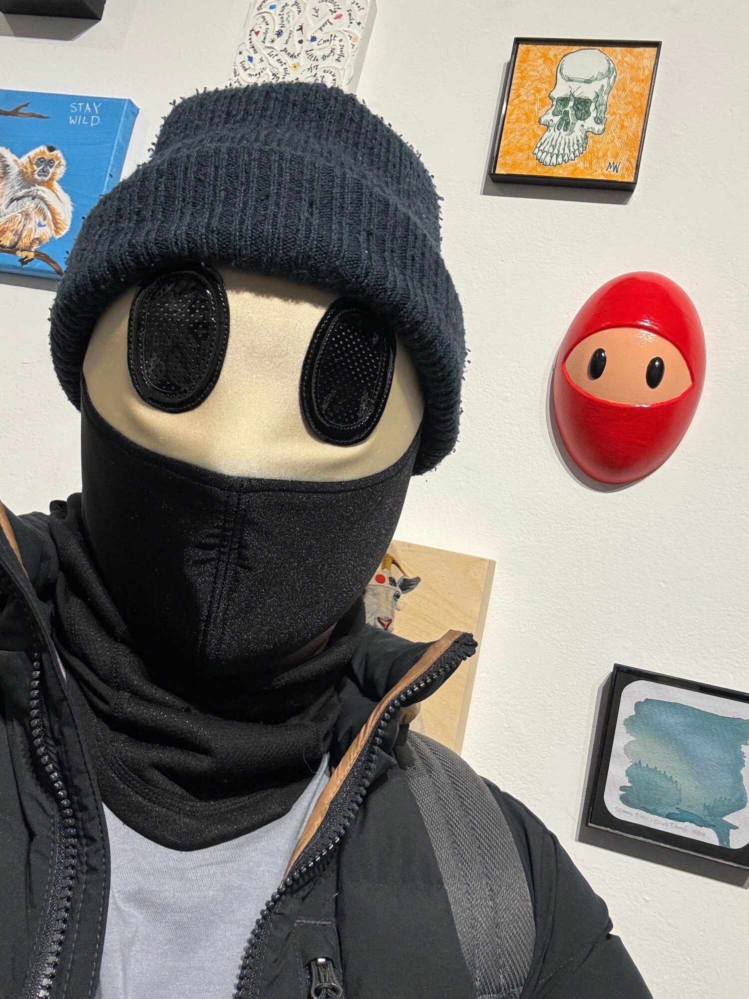 Person wearing a black mask, gray beanie, and gray jacket, with a backpack, in front of a wall with artwork and decorations.