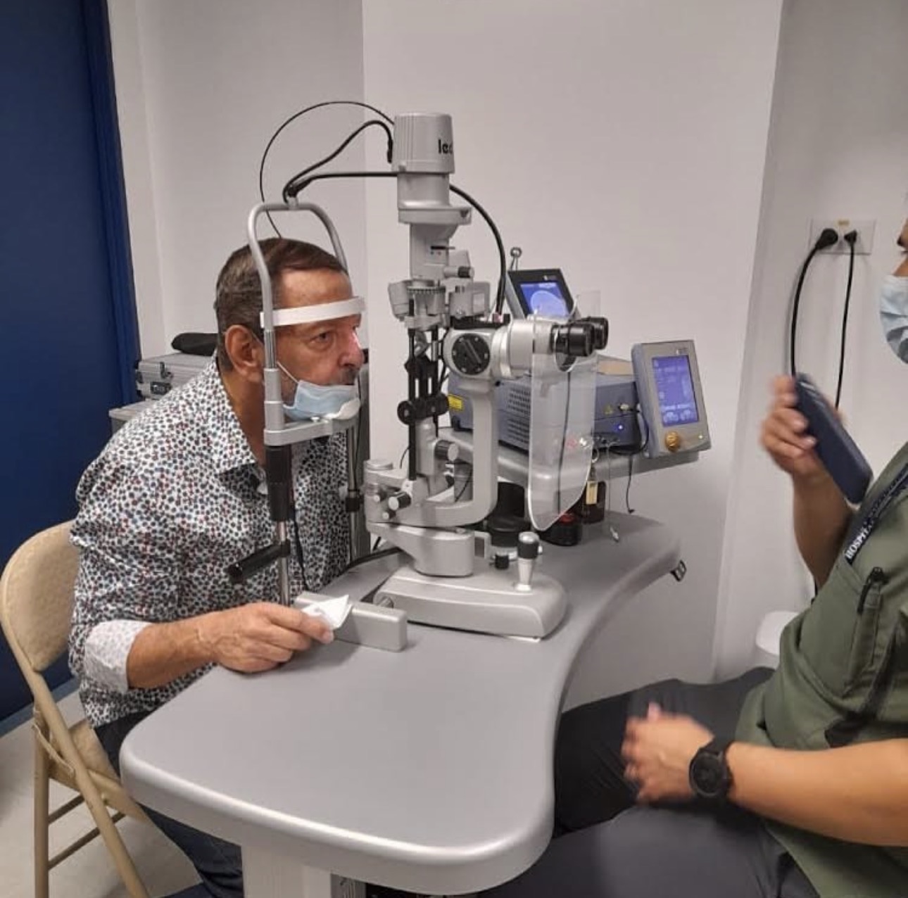 Man undergoing eye examination with a slit lamp, with another person holding a device nearby, in a clinical setting.