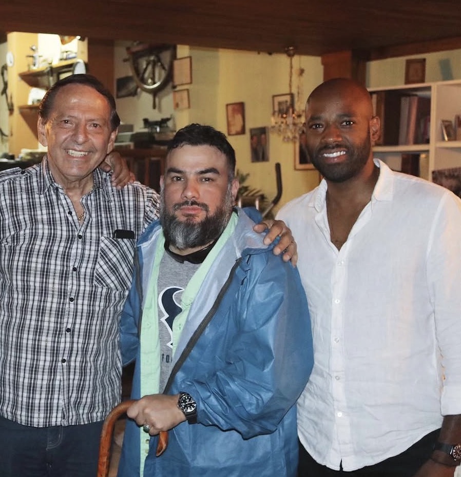 Three men standing indoors, smiling, with bookshelves and pictures in the background.