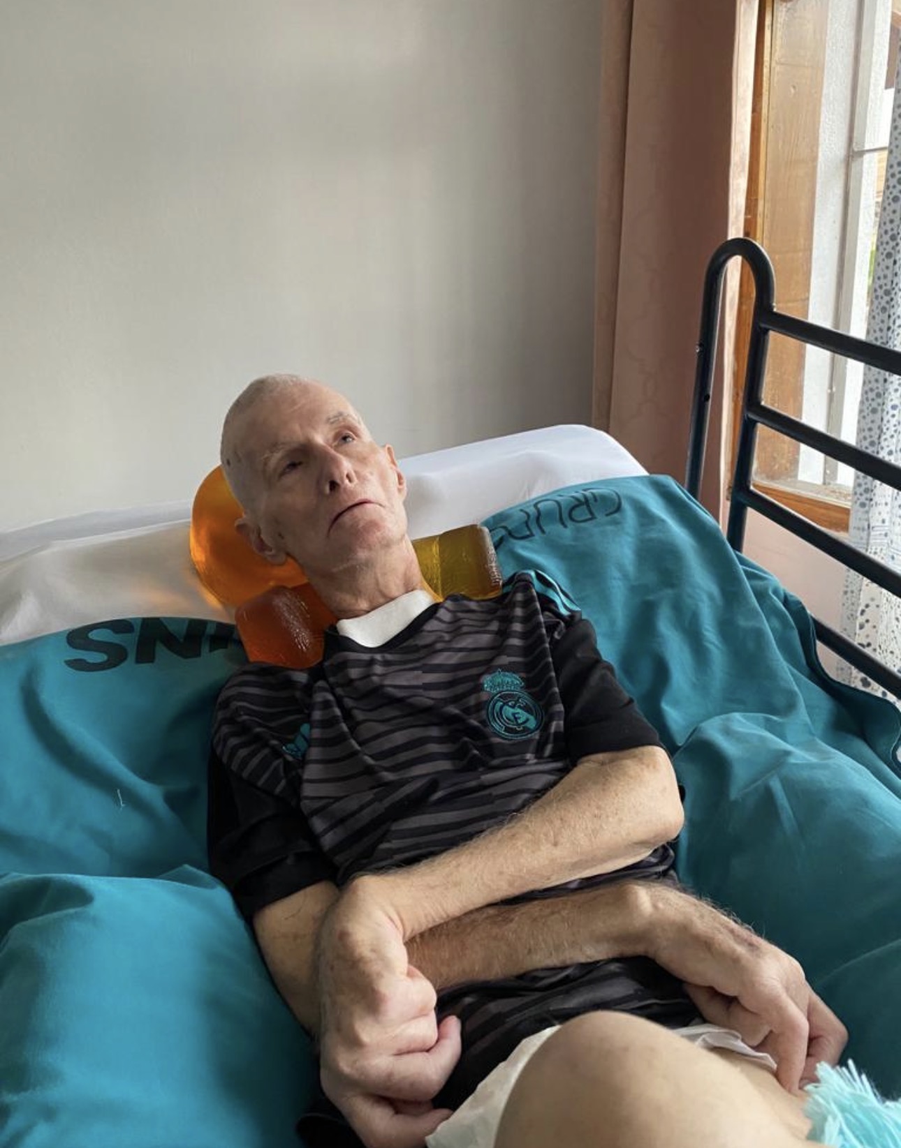 Elderly man lying in bed with head supported by an orange pillow, wearing a black striped shirt, in a room with curtains.