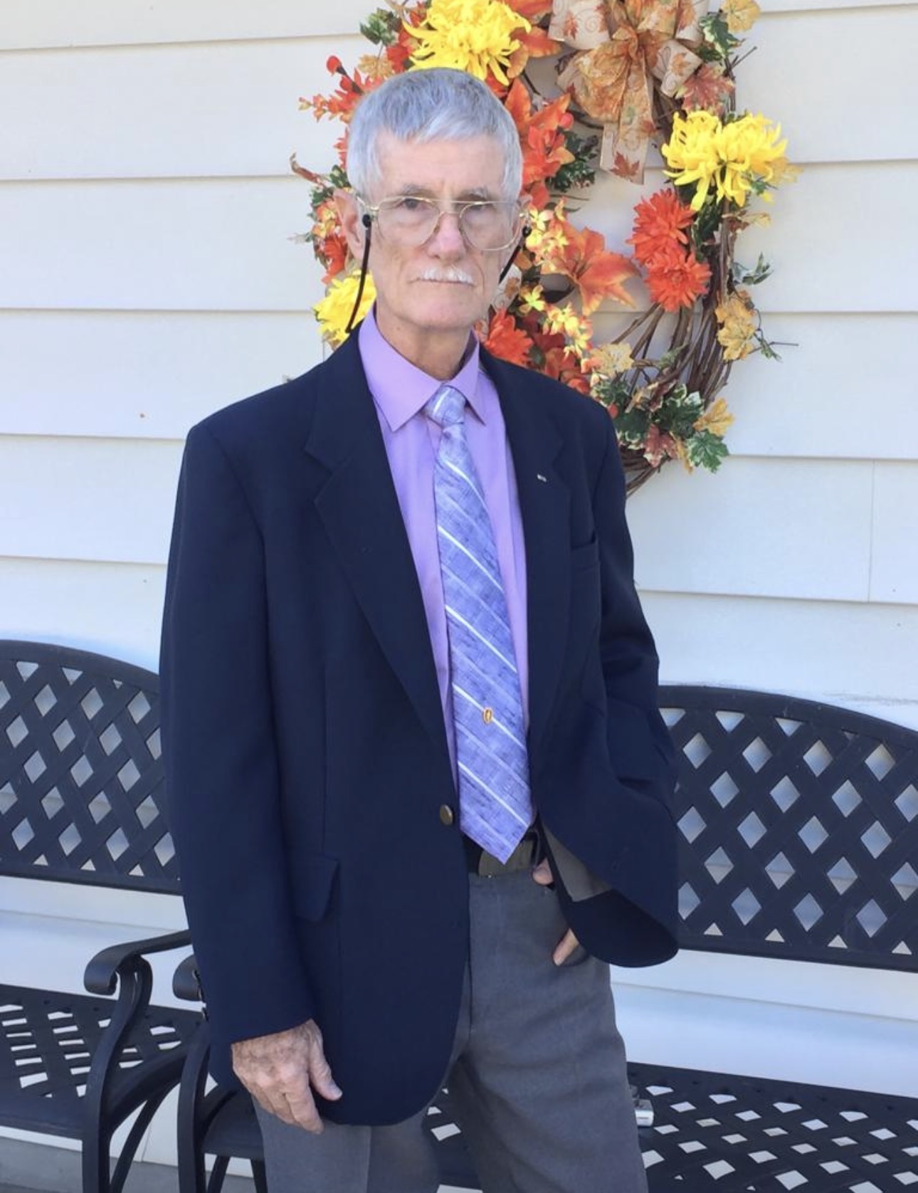 Elderly person with short gray hair and glasses, wearing a dark blazer, purple shirt, and patterned tie, standing outdoors in front of a floral wreath and a bench.
