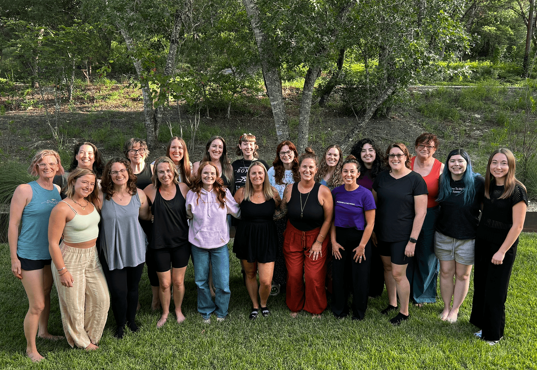 Group of 17 women standing outdoors on grass with trees in background, smiling for a photo.