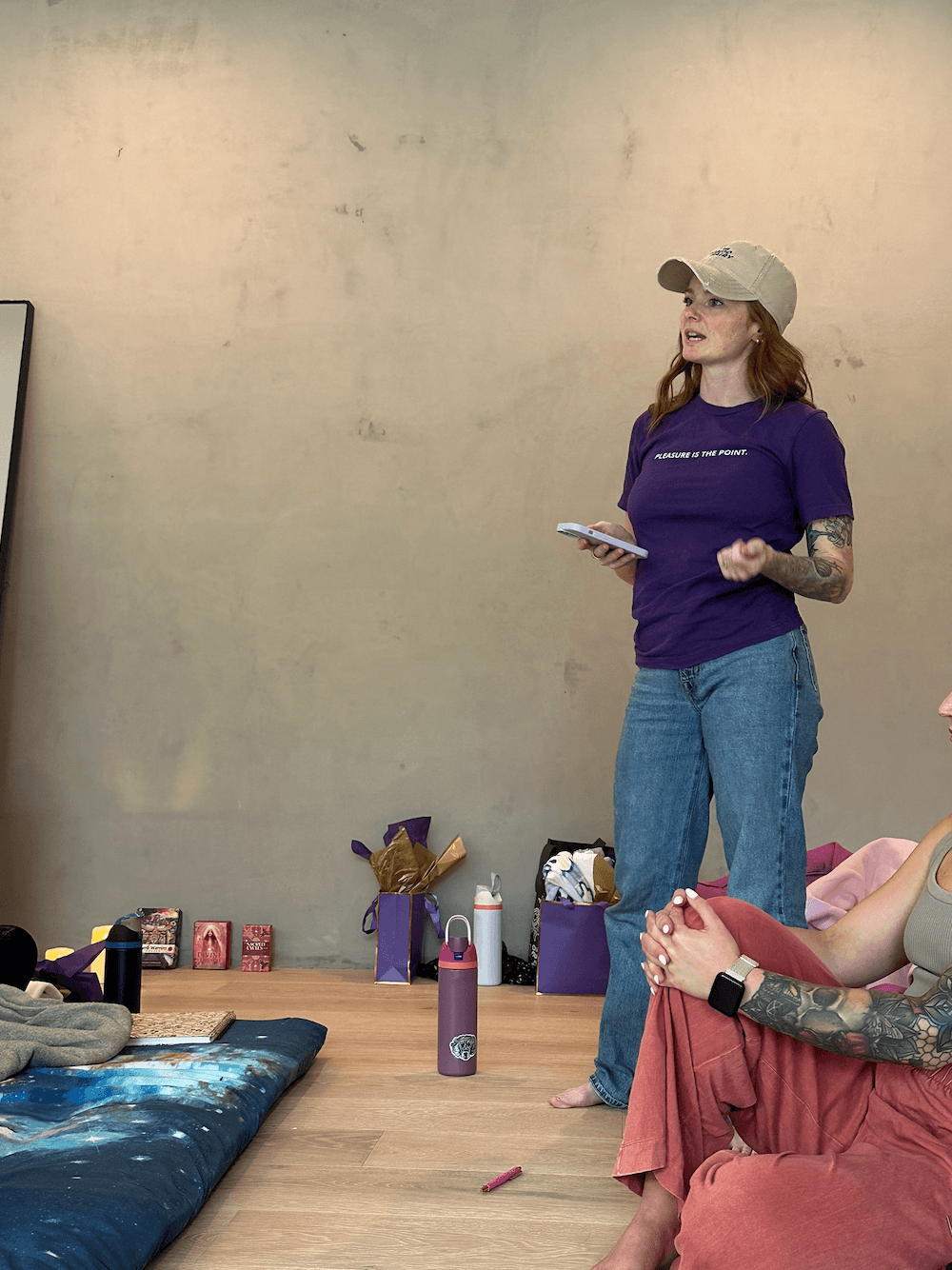 Woman with a cap and purple shirt stands holding a notebook, tattoos visible on her arm, in a room with a mattress and boxes.