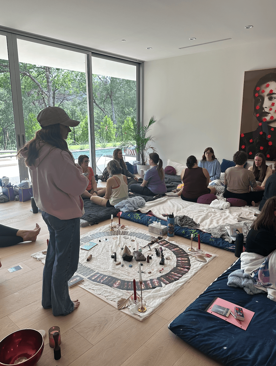 Group of people sitting on cushions and blankets in a room with large window, participating in a circle activity, with a woman standing nearby.