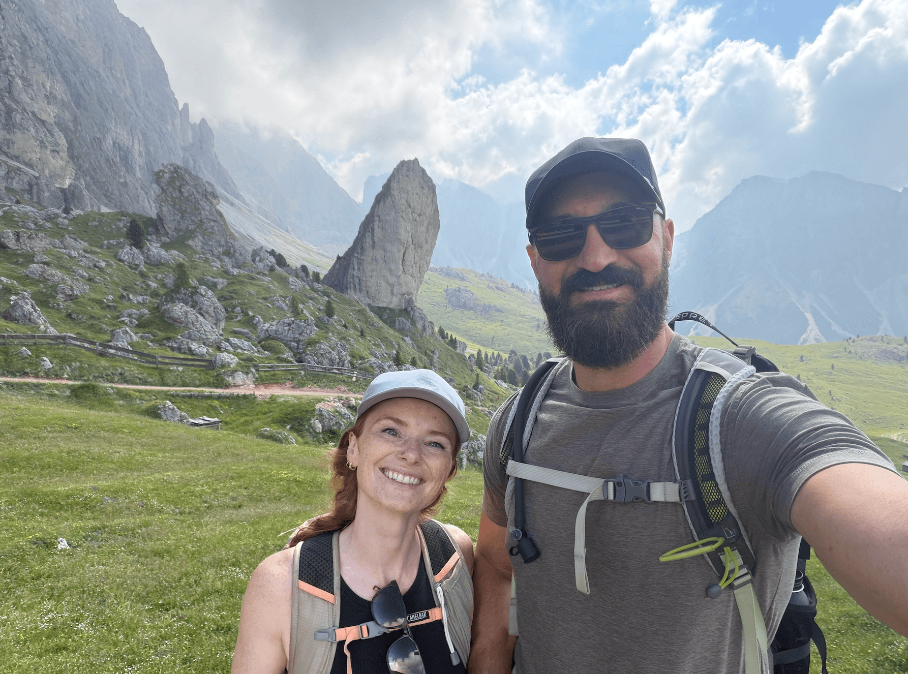 Two people outdoors in a mountainous landscape, smiling, with green grass and rocky cliffs in the background.