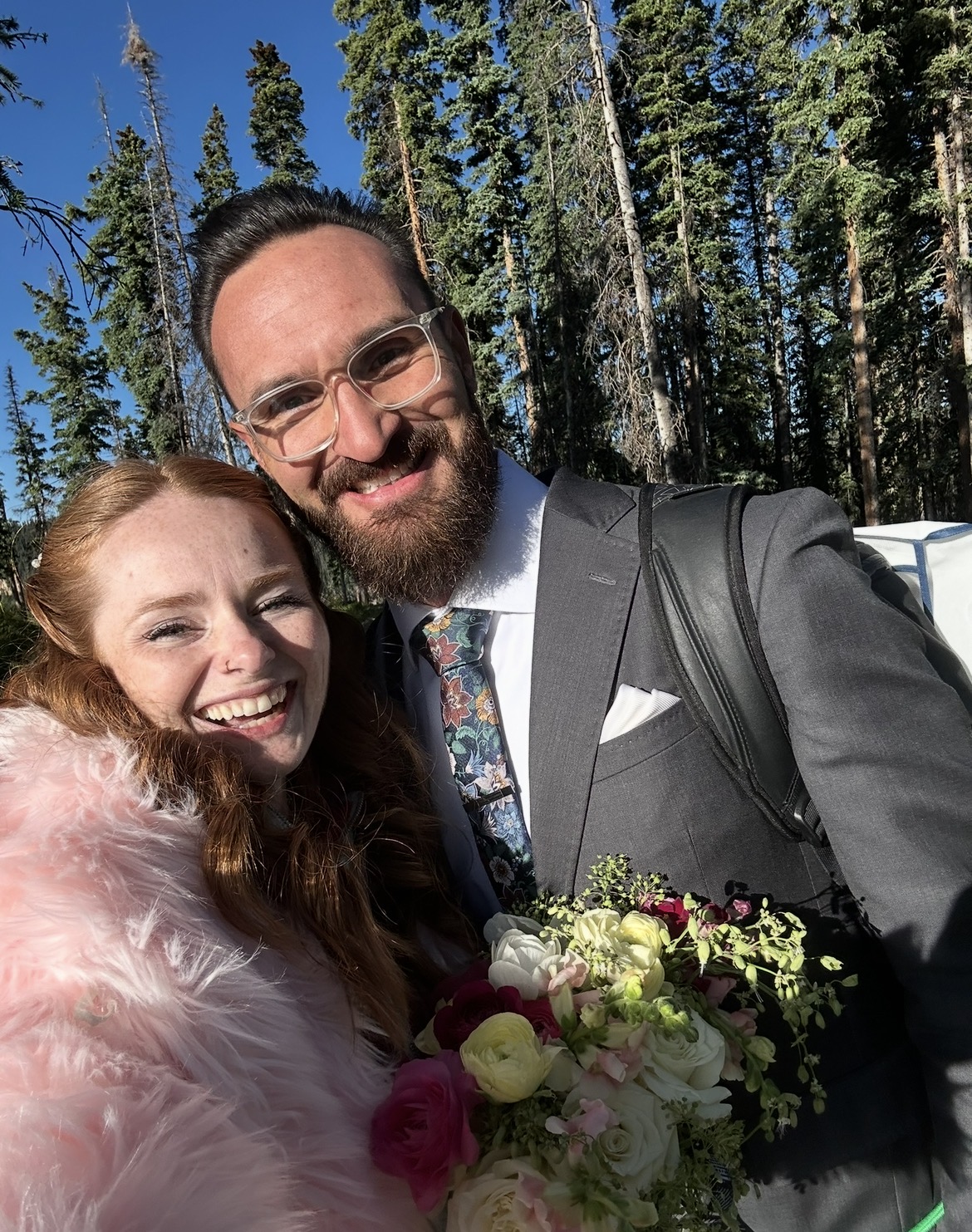 A smiling couple outdoors, the woman with red hair and pink fur coat, the man in a suit holding a bouquet of flowers.