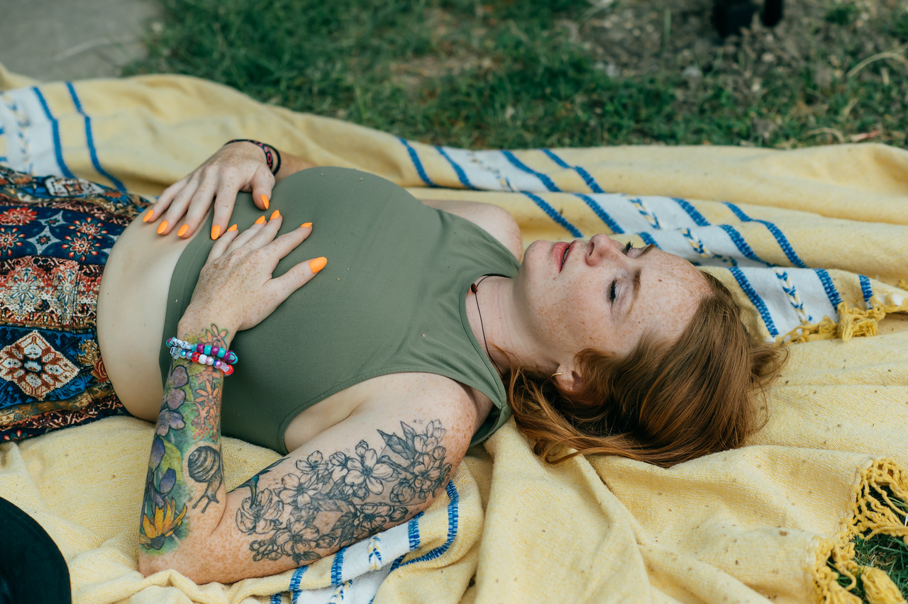 Woman with tattoos lying on a yellow blanket outdoors, eyes closed, wearing a green top and patterned skirt, with colorful bracelets.