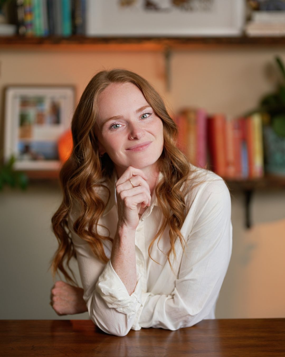 Young woman with long wavy hair, smiling, resting her chin on her hand, in a cozy room with bookshelves.