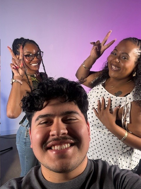 Three smiling people posing together, two women making peace signs, one man taking a selfie, colorful background.