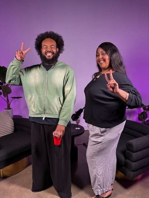 Two people smiling and making peace signs, standing in a room with purple lighting and modern furniture.