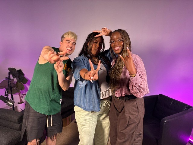Three people smiling and posing together in a colorful studio, with purple and pink lighting, making peace signs and gestures.
