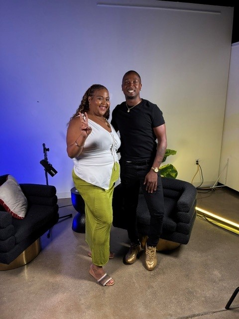 Two people standing together in a room with a blue and yellow gradient wall, black couches, and a potted plant.