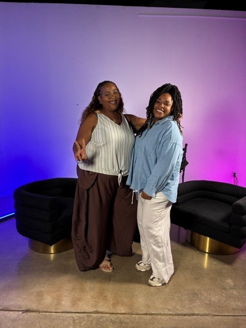 Two women stand together in front of a colorful gradient background, smiling and posing for the photo.