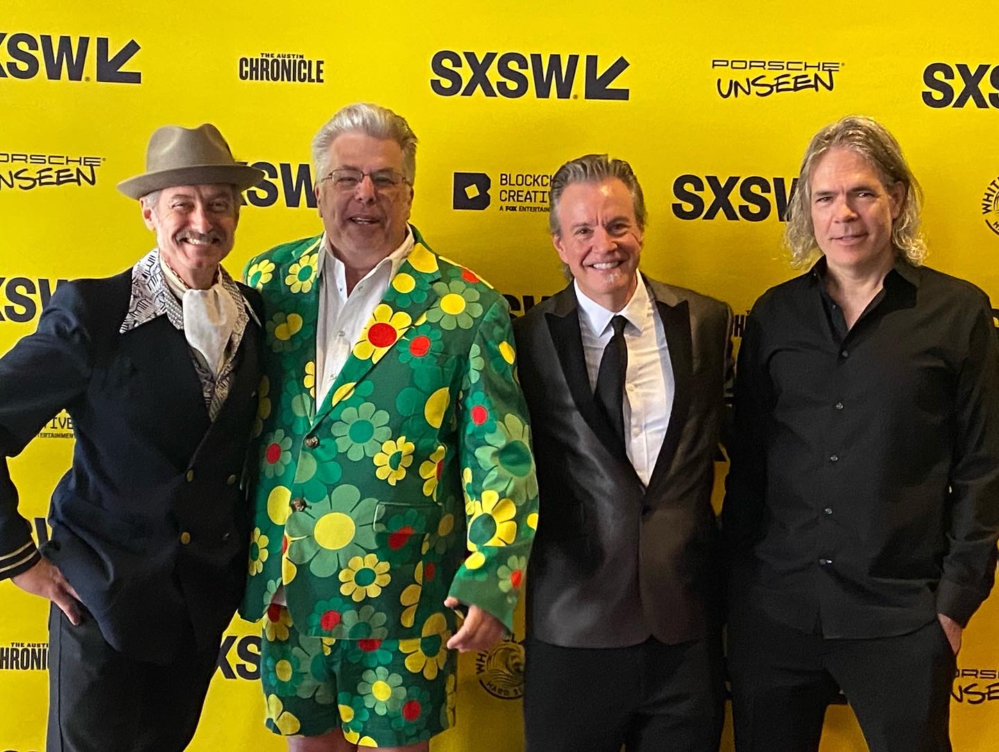 Four men standing together at SXSW event, smiling, with yellow background and event logos, one in a colorful floral suit.