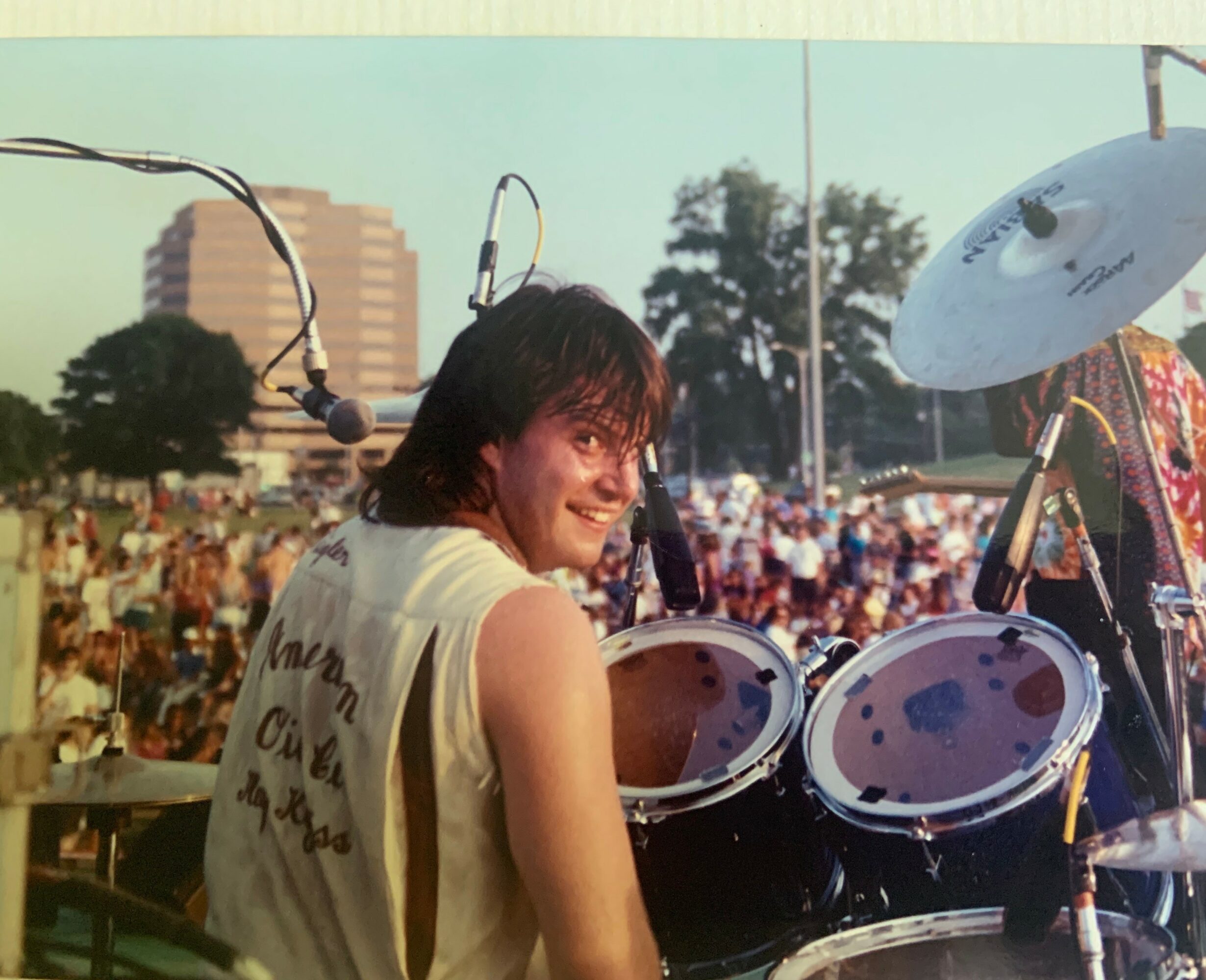 Person with dark hair smiling at a drum set outdoors during daytime, with a crowd and buildings in background.