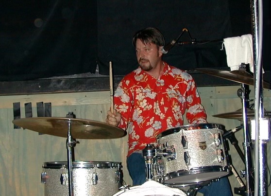 Man playing drums on stage, wearing a red floral shirt, with drum set and microphone visible.
