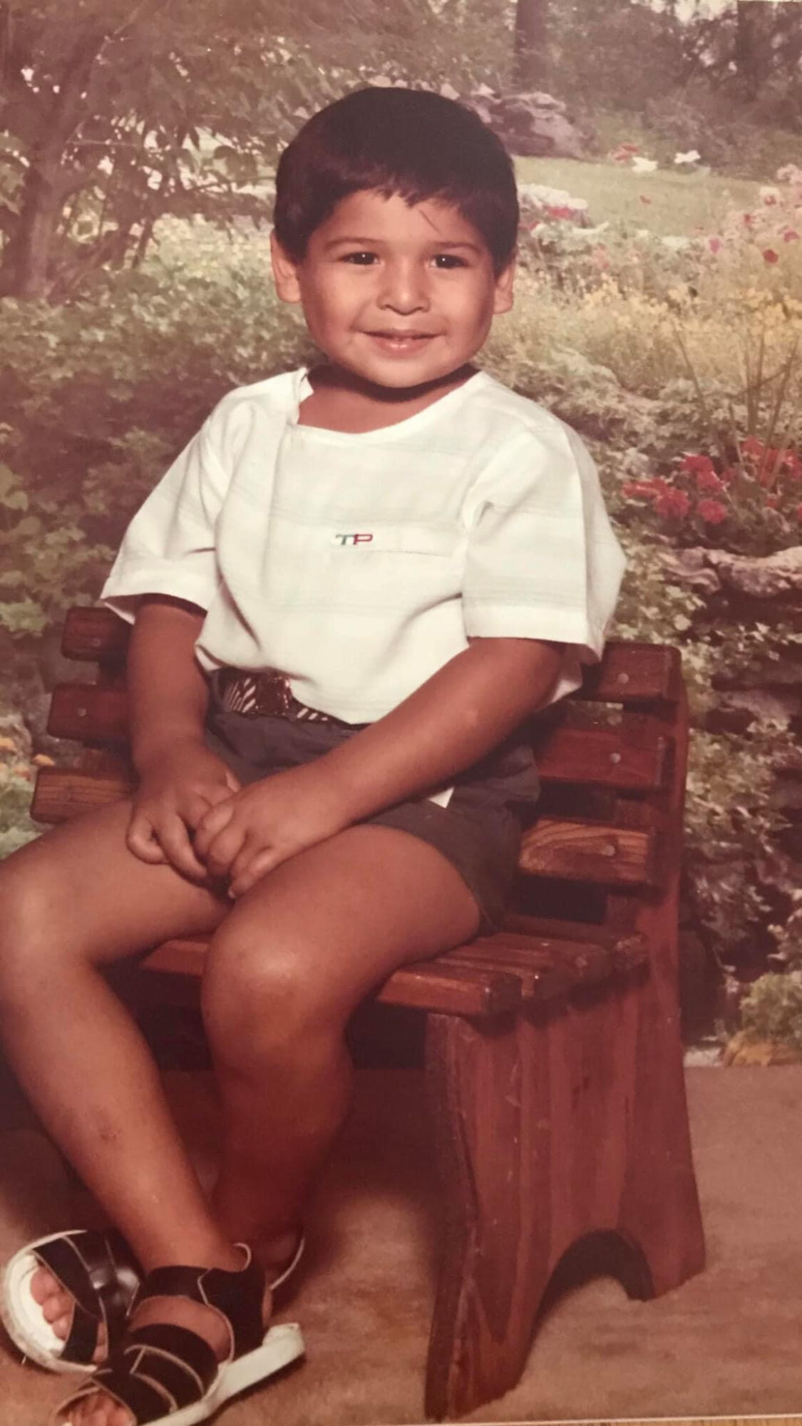 Young boy sitting on a small wooden bench outdoors, smiling, wearing a white shirt and shorts.