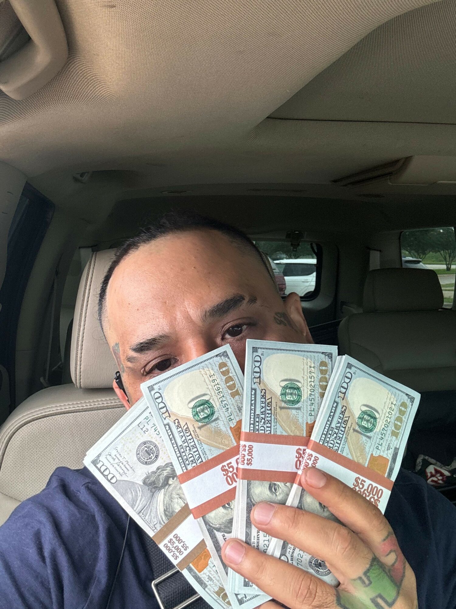 Person holding multiple hundred-dollar bills inside a vehicle, partially covering their face.