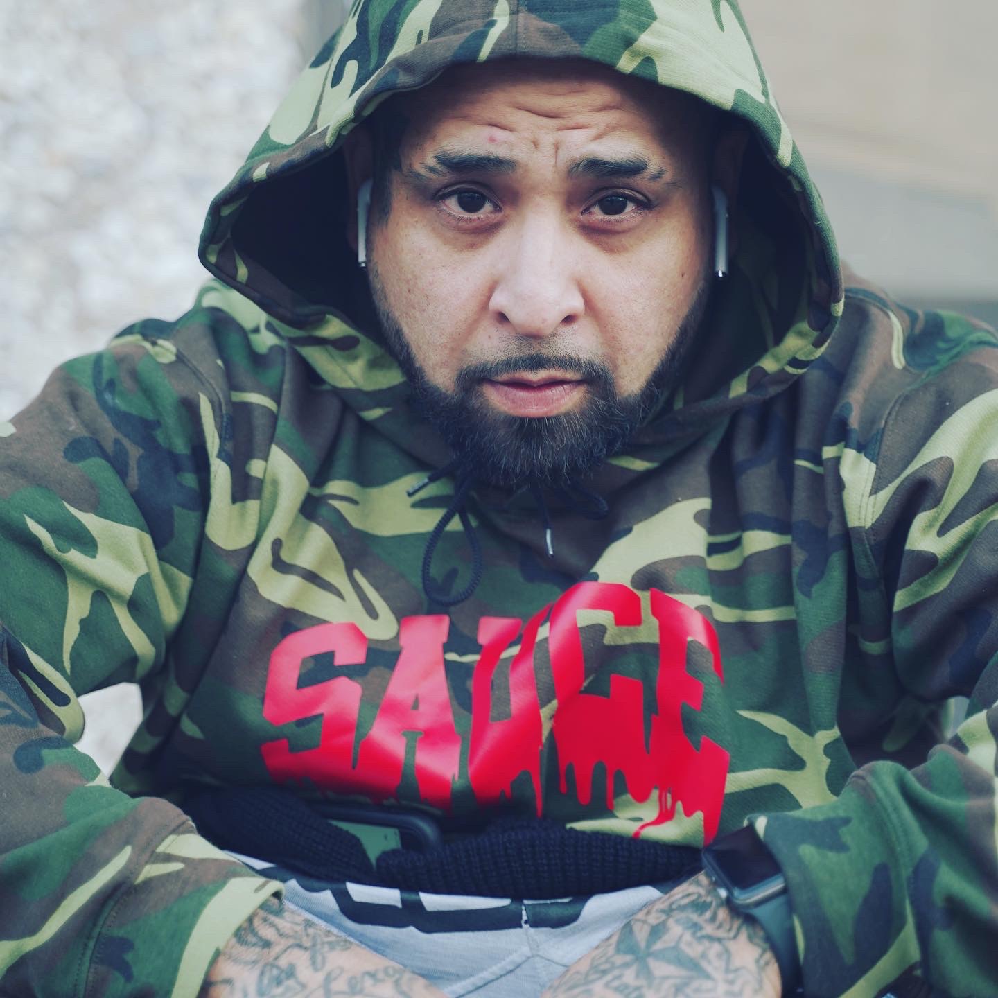 Man wearing camouflage hoodie and sweatshirt with red text, looking directly at camera, outdoors background