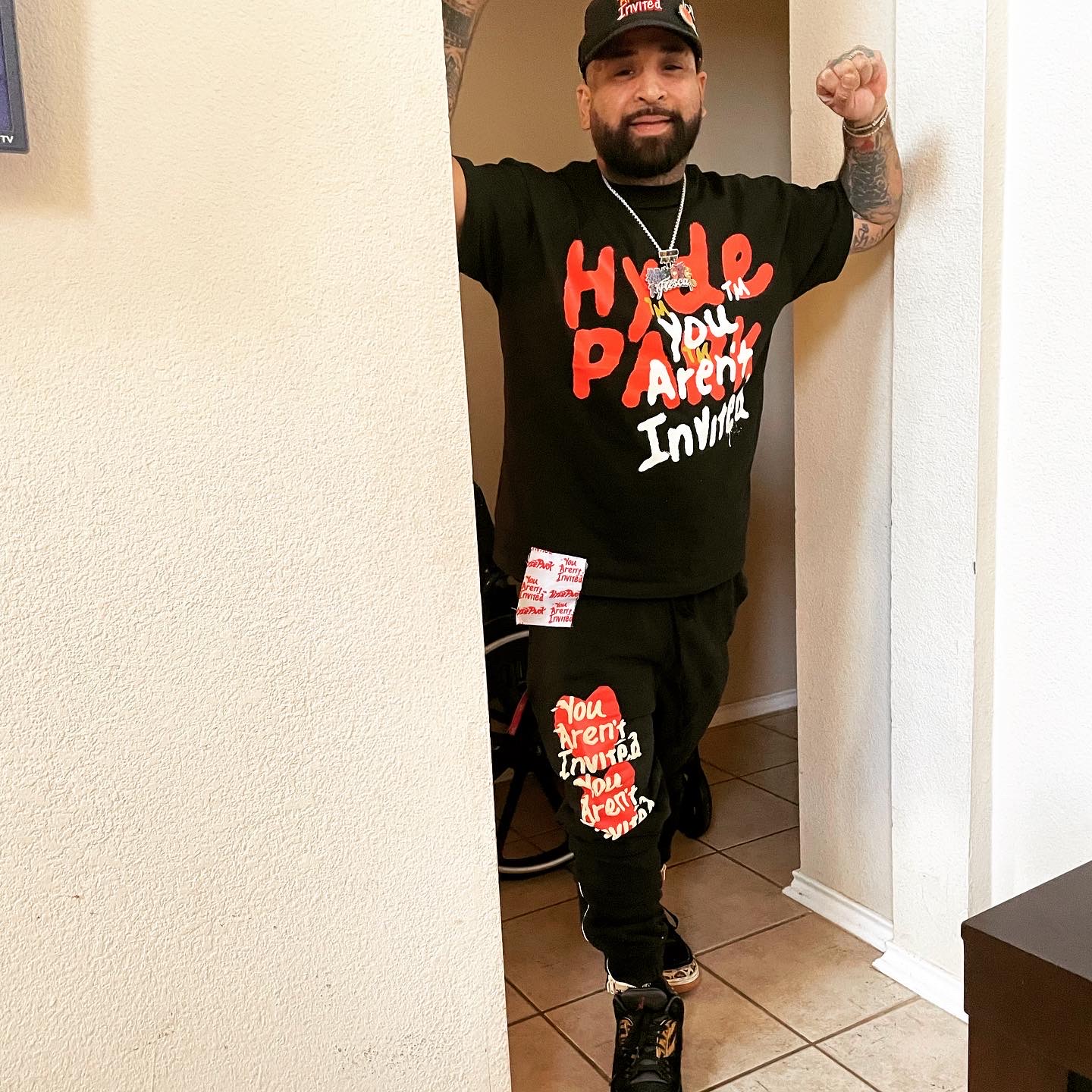 Man with beard and tattoos, wearing a black T-shirt and cap, stands in doorway with arms raised, smiling.