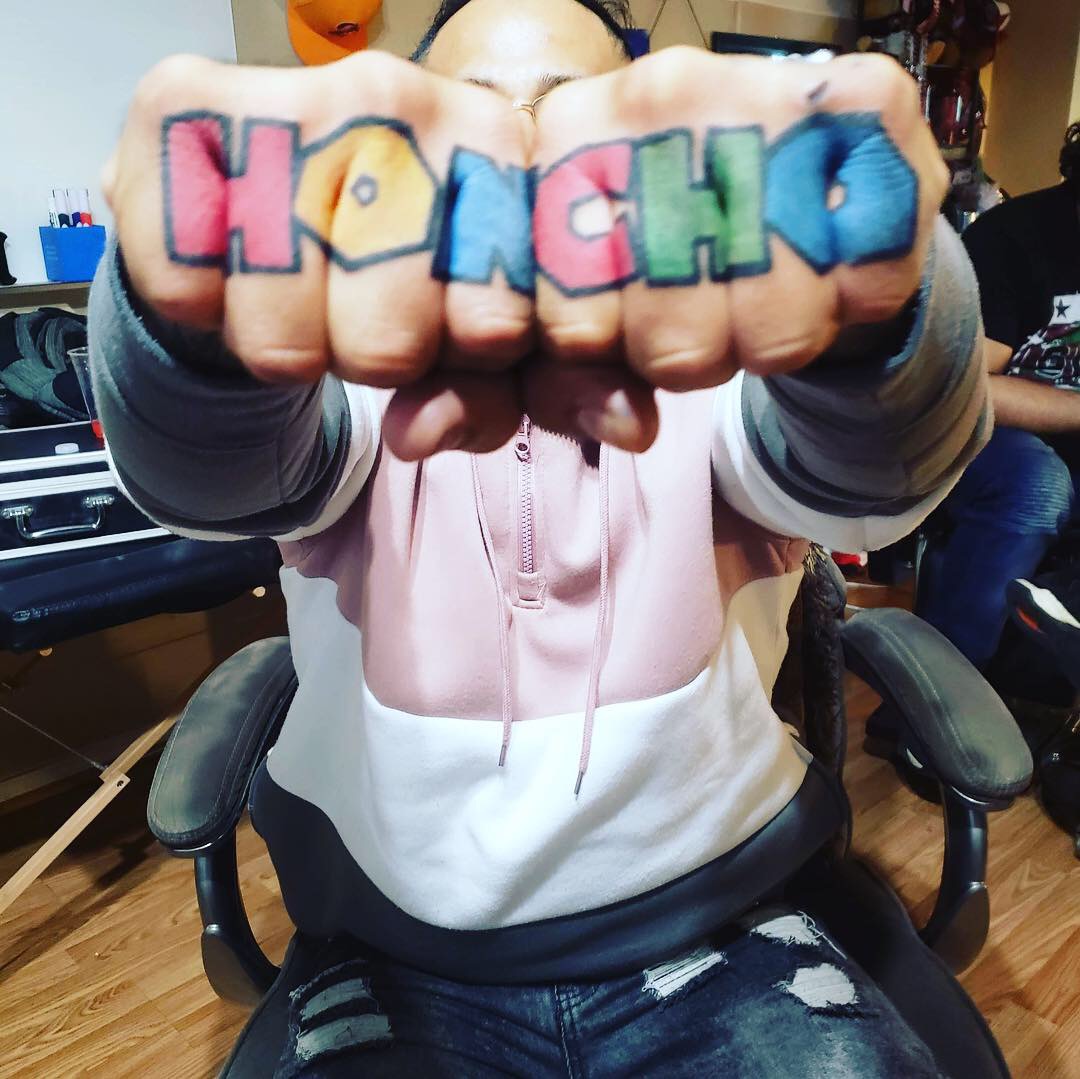 Person shows colorful tattooed fists with the word 'KOMCHU' in rainbow colors, in a room with chairs and people.
