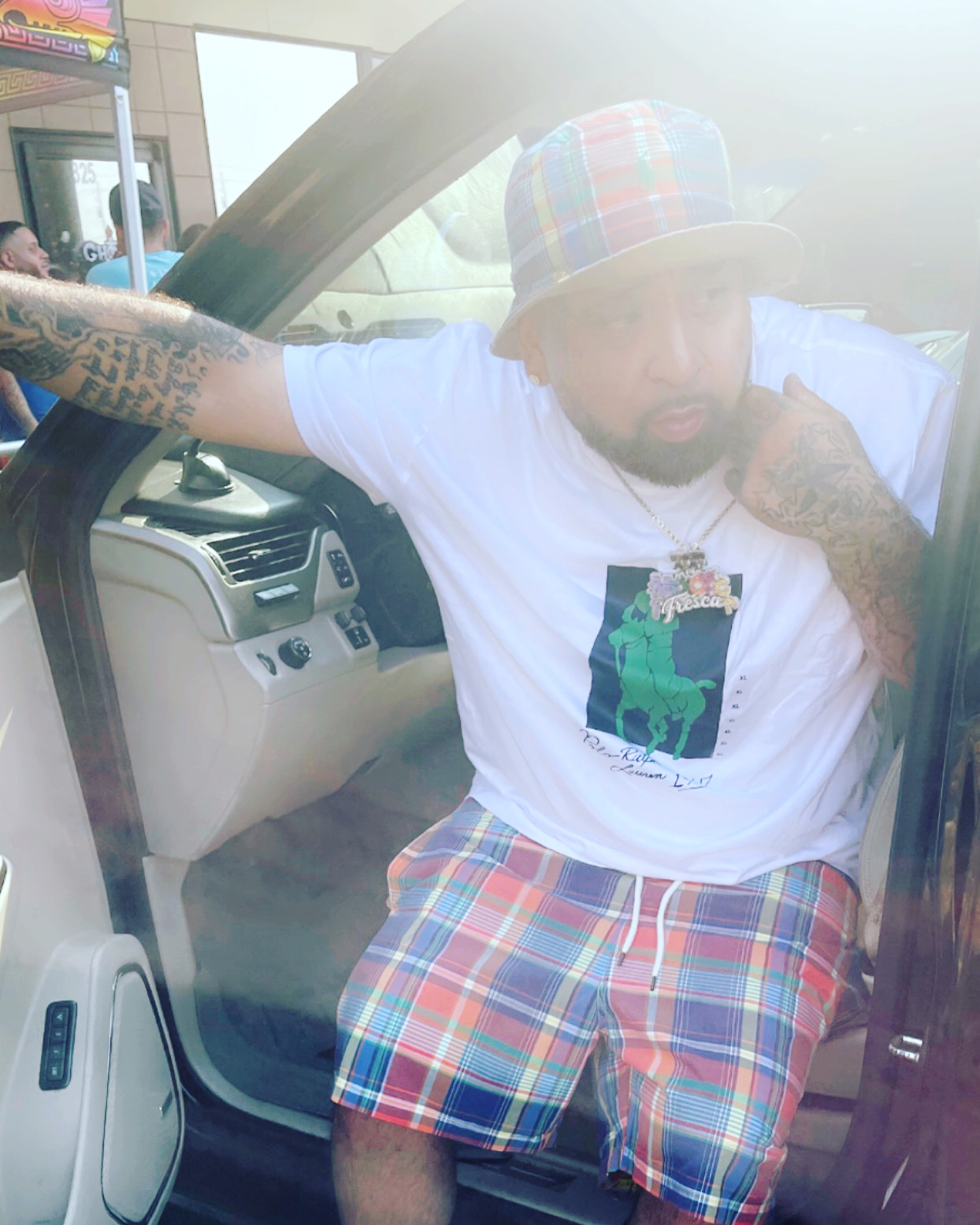 Man sitting in a car with arm resting on the door frame, wearing a colorful hat, white T-shirt, and plaid shorts.
