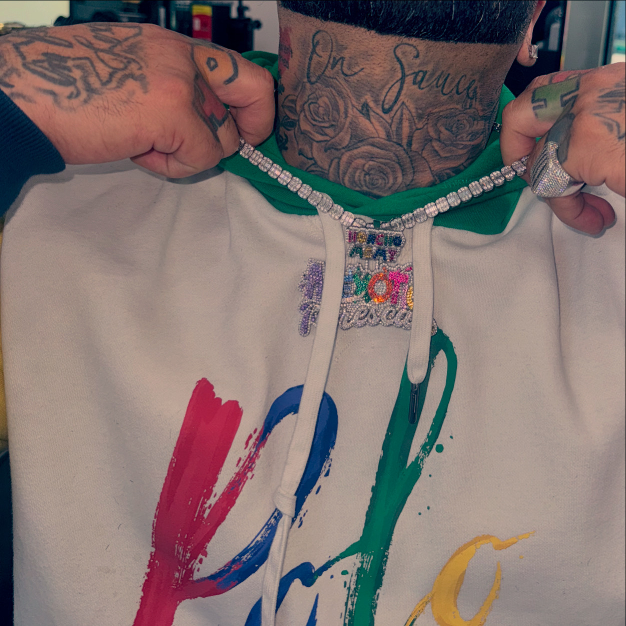 Person holding a bejeweled chain, wearing a hoodie with colorful paint strokes, tattoos visible on neck and hands.