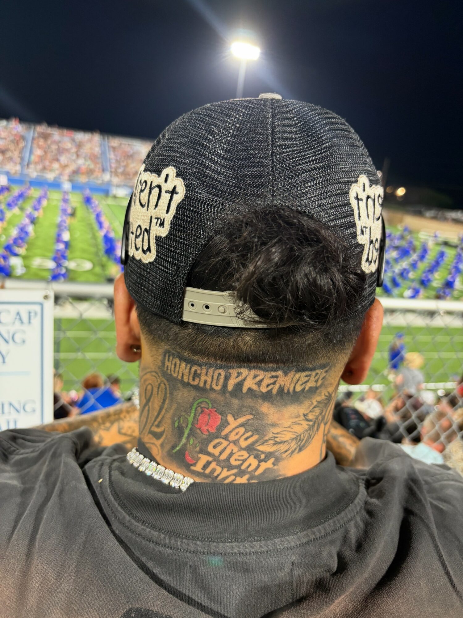 Person wearing a black cap with text, showing tattoos on the neck, at a stadium event at night.
