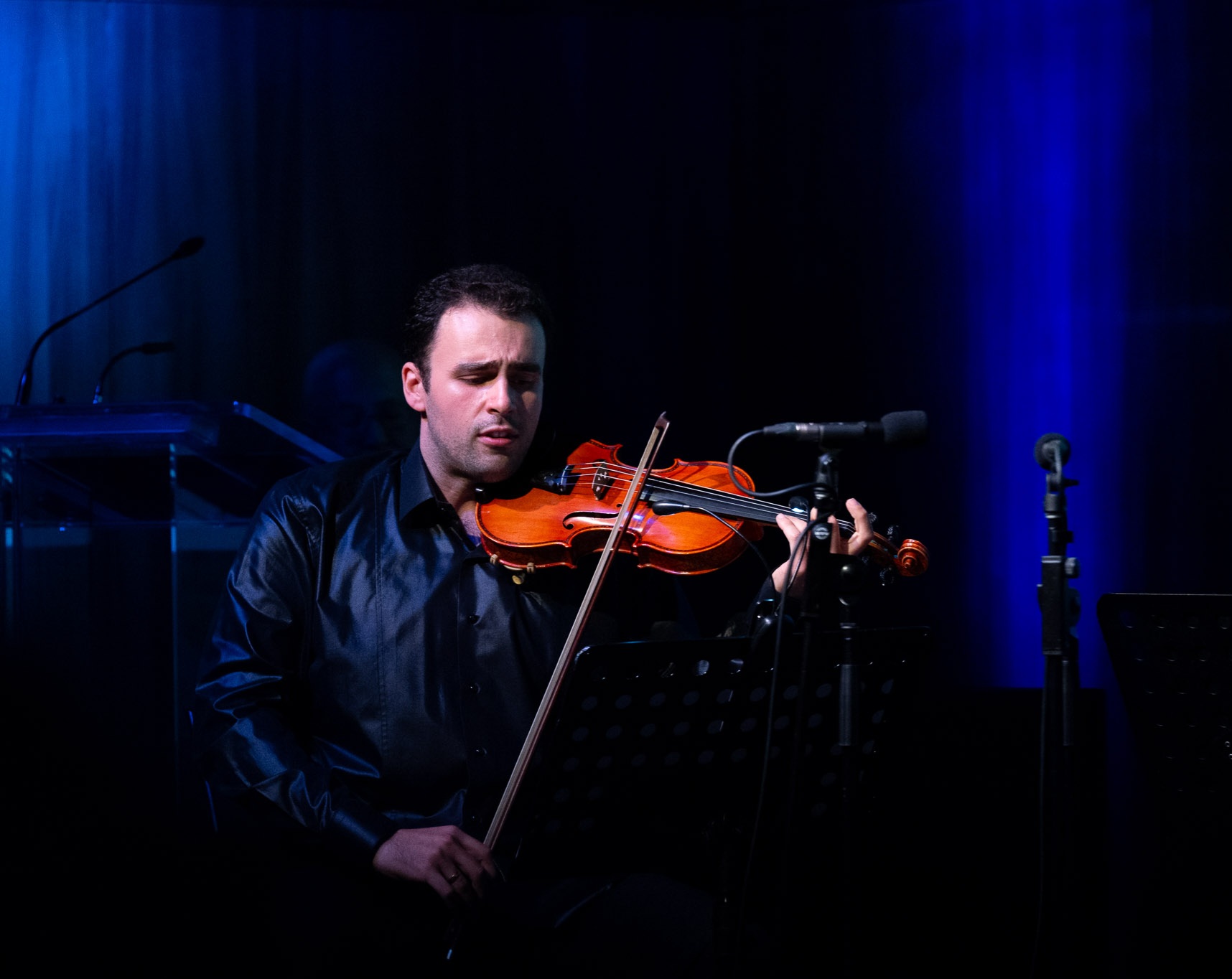 Man playing violin on stage with dark background and blue lighting, holding bow and looking focused.