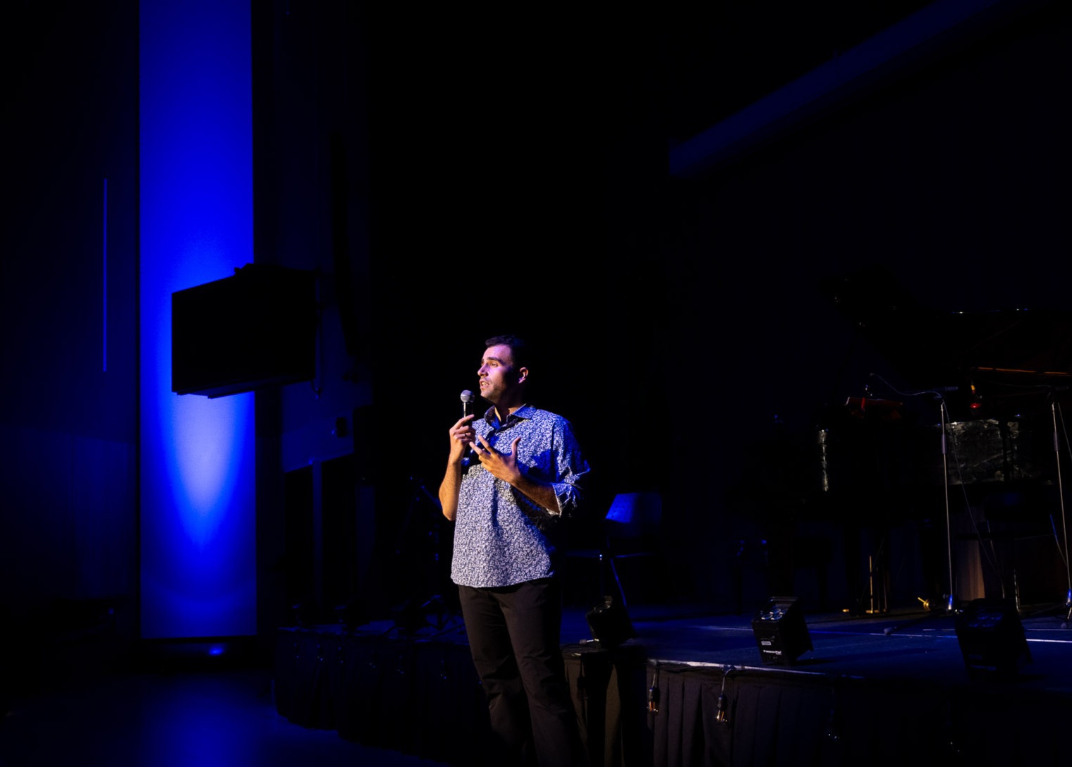 Person standing on stage holding a microphone, with blue lighting on the left side, dark background, and musical instruments nearby.