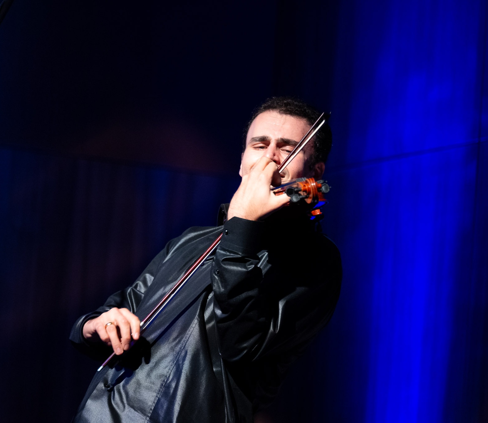 Person playing a violin with a blue background, eyes closed, wearing a black jacket.