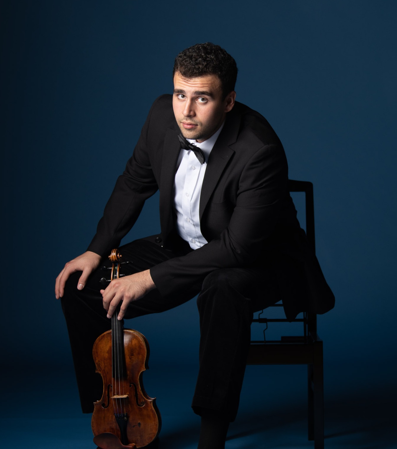 Man in tuxedo sitting on a chair with a violin, against a dark blue background.