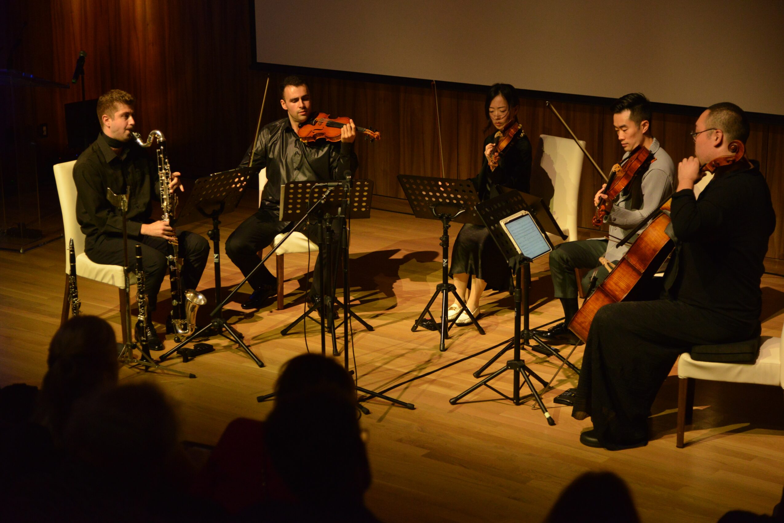 Five musicians perform on stage with string instruments, seated with music stands, in a wooden-paneled room.