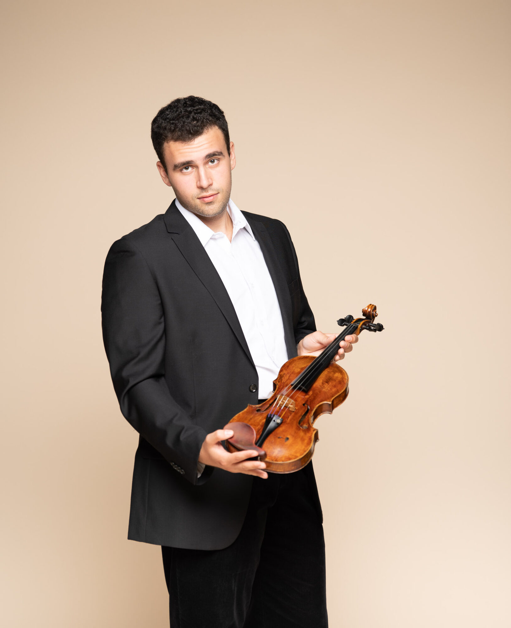 Young man in a black suit holding a violin against a beige background.