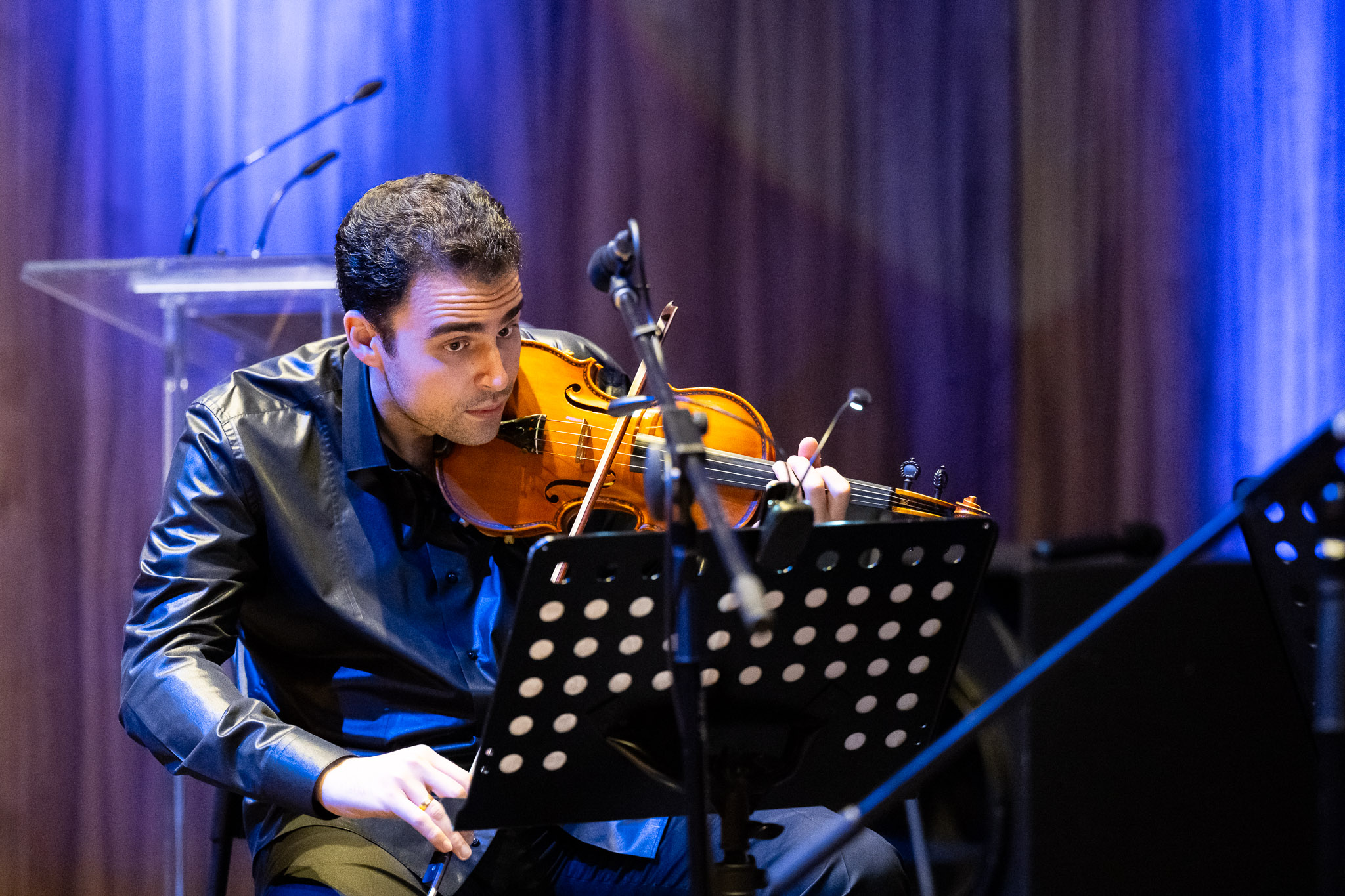 Person playing violin on stage with music stand, purple curtain background, stage lighting, and microphone, focused on music.