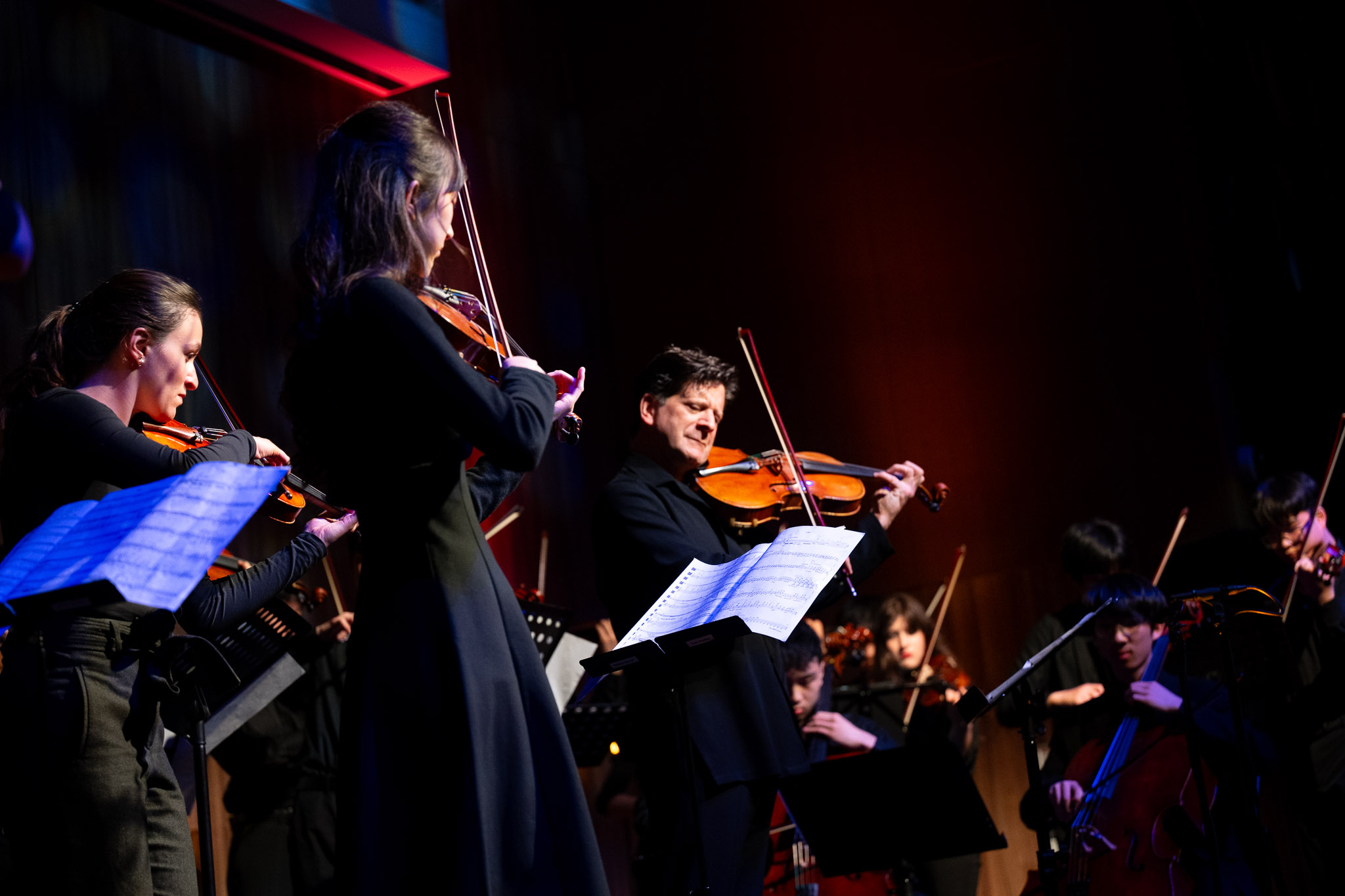 Group of young musicians playing violins on stage with music stands, in a concert setting.