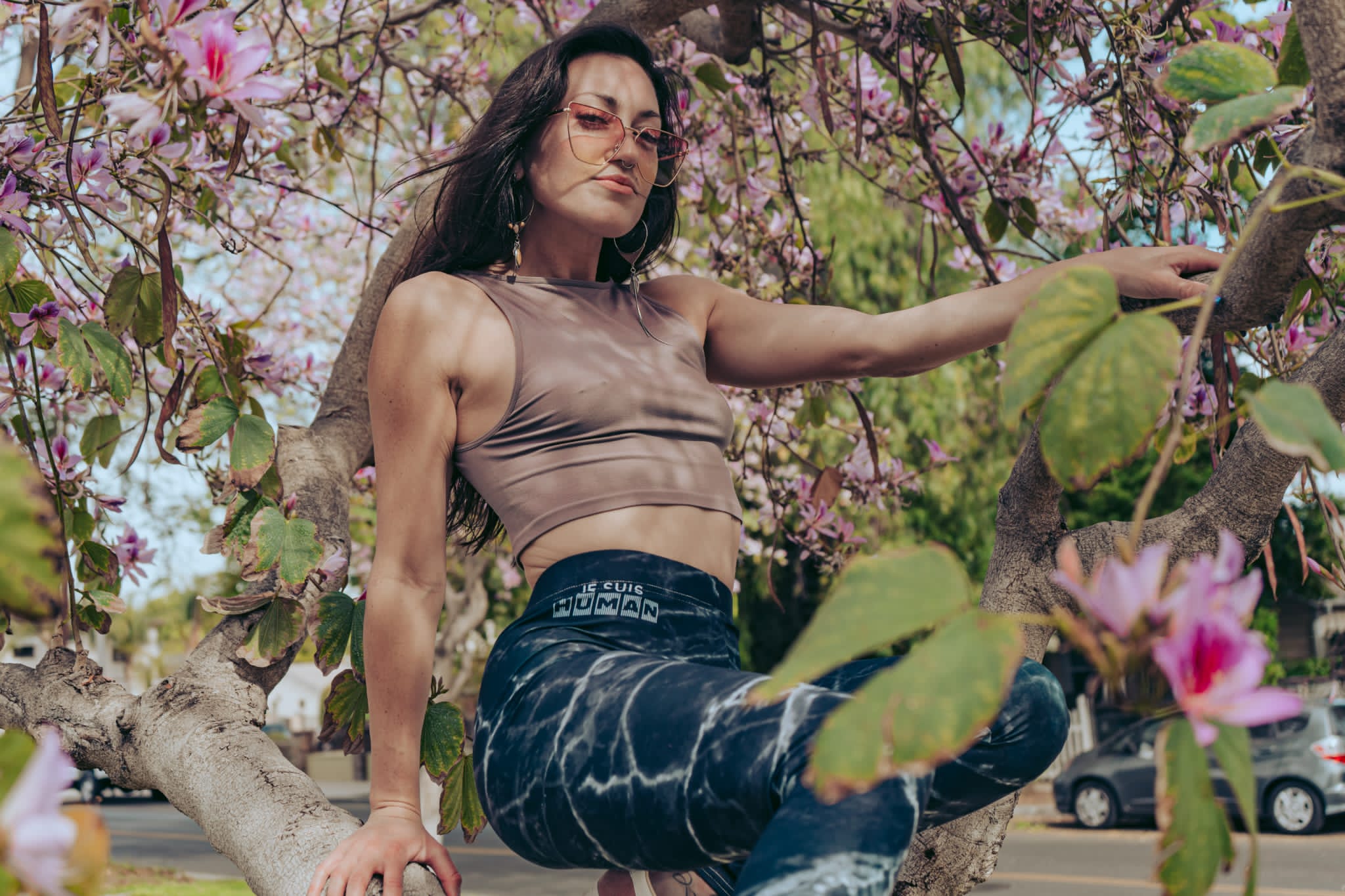 Woman sitting on a tree branch among pink blossoms, wearing a sleeveless top and patterned pants, with cars in the background.