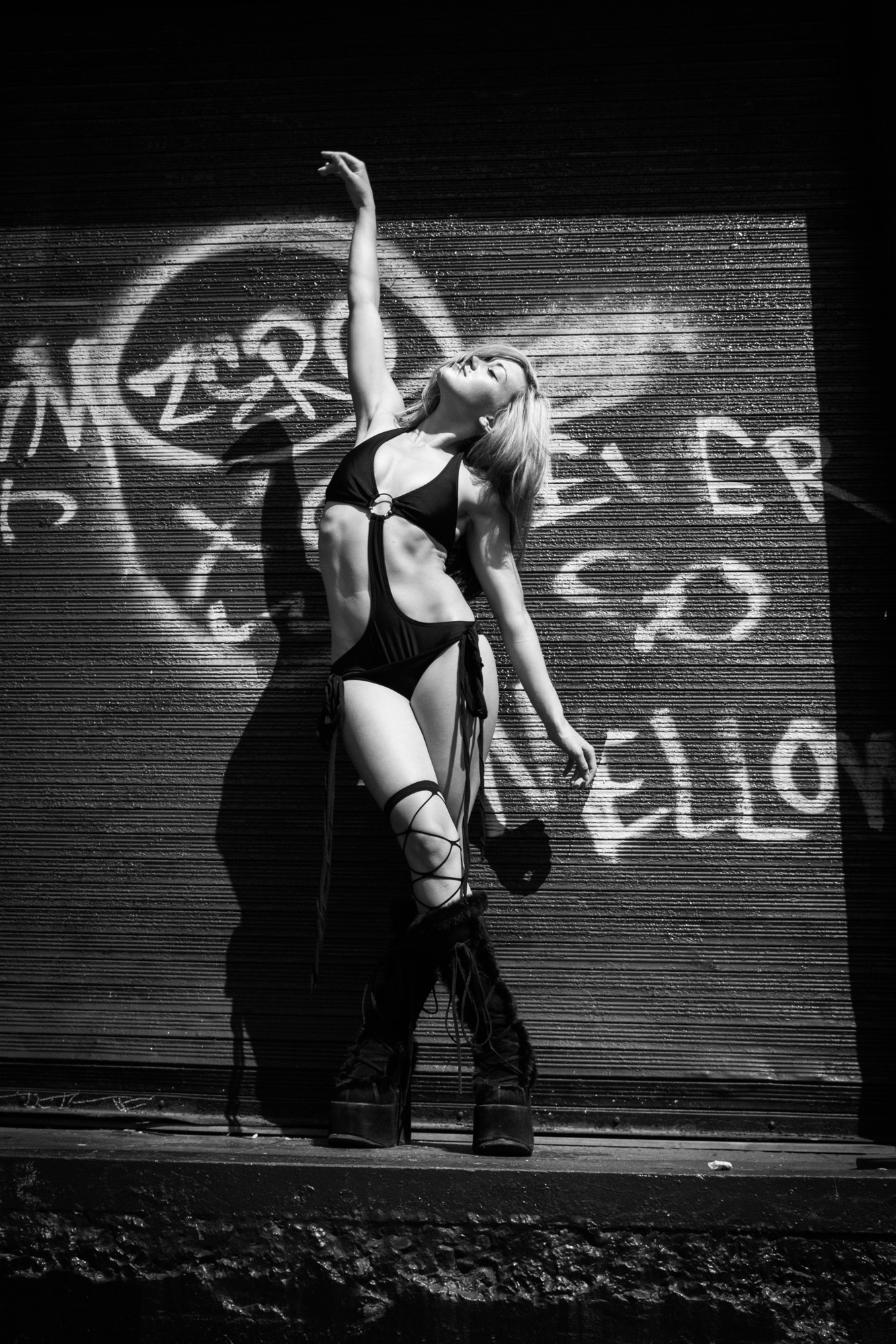 A woman in a black outfit poses with one arm raised, standing against a graffiti-covered wall, in black and white.
